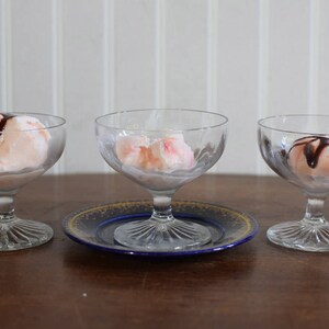 Puede incluir: Tres cuencos de postre de vidrio transparente con helado rosa y salsa de chocolate. Los cuencos est&aacute;n sobre una mesa de madera con un plato decorativo azul y dorado debajo de uno de los cuencos.