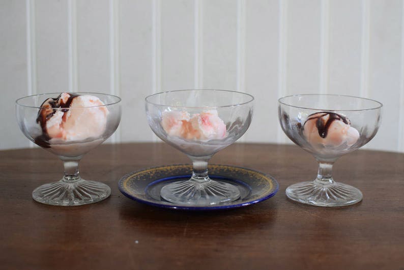 Puede incluir: Tres cuencos de postre de vidrio transparente con helado rosa y salsa de chocolate. Los cuencos est&aacute;n sobre una mesa de madera con un plato decorativo azul y dorado debajo de uno de los cuencos.