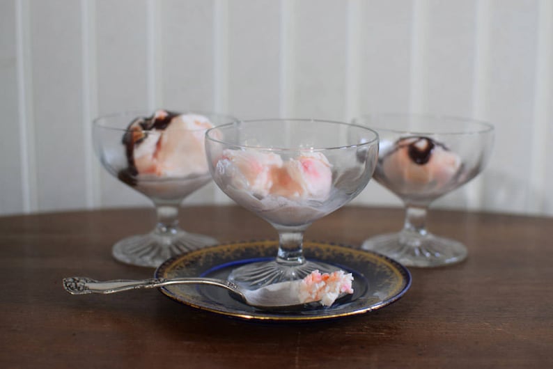 Puede incluir: Tres cuencos de postre de vidrio transparente llenos de helado rosa cubierto con salsa de chocolate. Una cuchara de plata con una bola de helado rosa encima se encuentra sobre un plato decorativo azul y dorado.