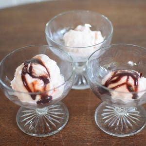 Puede incluir: Tres copas de postre de vidrio con helado rosa cubierto con salsa de chocolate.