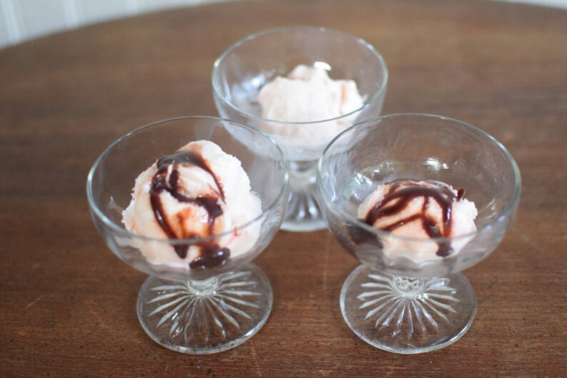 Puede incluir: Tres copas de postre de vidrio con helado rosa cubierto con salsa de chocolate.