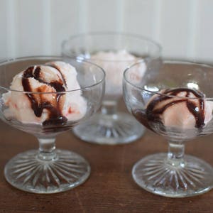 Puede incluir: Tres copas de postre de vidrio transparente con base con tallo, cada una llena con una bola de helado rosa cubierta con un chorrito de salsa de chocolate.