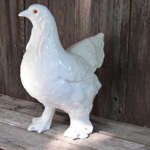 May include: A glossy white ceramic chicken figurine standing in profile. The sculpture features detailed molded feathers, a smooth curved body, and a distinct beak with a small orange eye. The surface has a reflective finish and stands upright.
