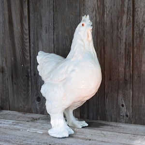 May include: A white ceramic chicken figurine standing in profile. The sculpture features detailed molded feathers, a prominent comb, and a wattle. The eyes are painted a distinct orange color, providing a contrast to the glossy white finish of the bird.