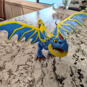 May include: A blue and yellow toy dragon with large wings. The dragon has a smiling face with yellow teeth and orange horns. The wings are blue with yellow accents. The dragon is posed on a speckled countertop.