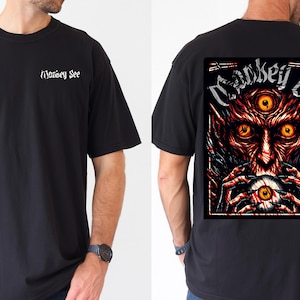 May include: Black t-shirt with the text "Money See" on the front and a graphic on the back. The graphic features a red and orange demon with three eyes holding an eyeball. The shirt is a short-sleeved, crew neck style.