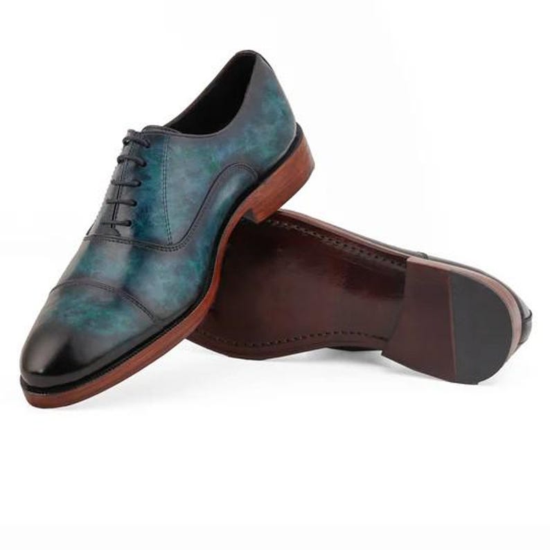 May include: A pair of oxford shoes featuring a teal and black marbled leather upper with a cap toe design, black laces, and a stacked wooden heel with a dark brown sole. The shoes are positioned to show both the side profile and the bottom sole.