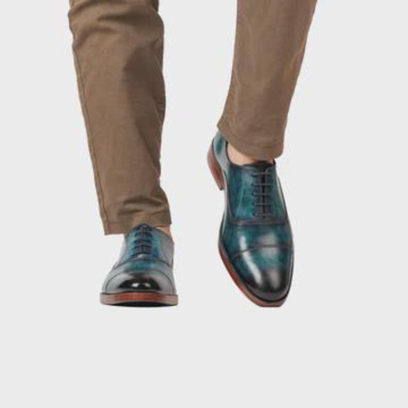 May include: A pair of teal and black leather oxford shoes with a cap toe design, black laces, and a brown sole, worn with brown trousers. The shoes feature a polished finish with a gradient color transition from the dark toe to the teal body.
