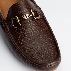 May include: A dark brown loafer featuring a textured, reptilian-inspired embossed pattern on the vamp. The shoe is accented with a polished gold-tone metal horsebit hardware piece that prominently displays a central D logo emblem.