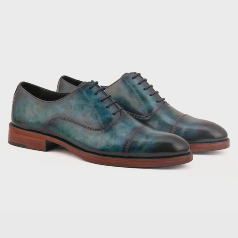 May include: A pair of oxford shoes featuring a cap toe design with a burnished teal and black gradient finish. The shoes have thin black laces, a low heel, and a stacked brown wooden sole, showcasing a polished leather construction throughout.