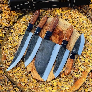 May include: A set of five knives with dark, textured metal blades and polished brown wood handles secured with silver rivets. The knives are arranged on a circular wooden block, with a black leather carrying case positioned above them.