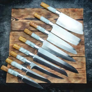 May include: An overhead shot of a collection of handcrafted kitchen knives. The knives have silver blades, light blue and white handles, and wooden accents. They are arranged on a wooden cutting board.