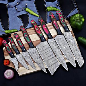 May include: A set of seven kitchen knives with patterned Damascus steel blades and multi-colored red and green wood handles, arranged on a wooden cutting board alongside fresh green peppers, a red onion, and a red tomato.