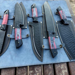 May include: A set of five kitchen knives with black hammered blades and handles featuring red and grey wood grain patterns. Each knife is paired with a matching black sheath, and the entire collection is arranged on a light blue leather roll.