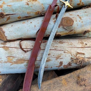 May include: A curved sword with a patterned Damascus steel blade, a gold-colored metal crossguard, and a black handle with white pins. The sword is accompanied by a brown leather scabbard featuring a textured finish and an adjustable strap.