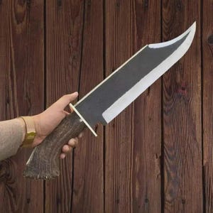 May include: A large fixed-blade knife featuring a dark, textured metal blade with a polished silver edge. The handle is crafted from a brown, rugged antler material, separated from the blade by a thin, flat brass-colored metal guard.