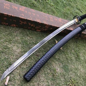 May include: A Japanese katana sword with a silver blade, black handle, and a black scabbard with a woven pattern. The sword rests on a brown box with Japanese characters. The items are on a grassy surface.