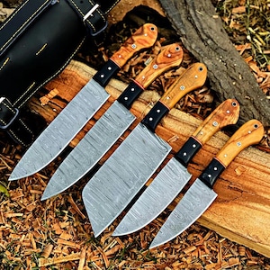 May include: A set of five knives with Damascus steel blades featuring a wavy pattern, black bolsters, and light brown wooden handles with two rivets. The knives are arranged in a row, showcasing varying blade shapes and sizes for culinary use.