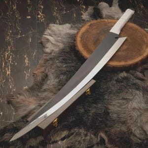 May include: A large, silver-bladed sword with a white-wrapped handle. The blade is angled and rests on a wooden stand with gold accents. The sword is displayed on a fur-like surface.