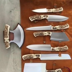 May include: A set of seven kitchen knives and a crescent-shaped blade with textured antler handles and polished steel blades, arranged on a brown leather roll-up storage case with brass snap fasteners and visible stitching details.