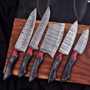 May include: A set of six kitchen knives with patterned steel blades and wooden handles. The handles are a combination of red, black, and green, with brass rivets. The knives are arranged on a wooden cutting board.