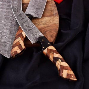 May include: Two kitchen knives featuring Damascus steel blades with a wavy, layered pattern. The handles are crafted from light and dark wood pieces arranged in a herringbone pattern, secured with brass pins and a central decorative mosaic rivet.
