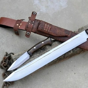 May include: Two knives with wooden handles and silver blades rest on a wooden surface. A brown leather sheath with a belt loop and decorative stitching is also present. A chain is visible in the lower left corner.