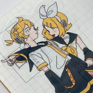 May include: A transparent sticker featuring two anime-style characters with yellow hair and black and yellow outfits. The characters are in a close embrace, with one pointing. The sticker is on a grid-lined surface.