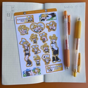 May include: An open notebook with a sticker sheet featuring cartoon characters and the text "KAGAMINE RIN" and "KAGAMINE LEN." Two orange and yellow pens are also visible. The notebook is open to a blank page.