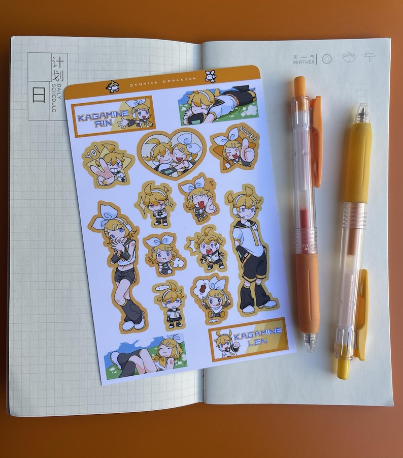 May include: An open notebook with a sticker sheet featuring cartoon characters and the text "KAGAMINE RIN" and "KAGAMINE LEN." Two orange and yellow pens are also visible. The notebook is open to a blank page.
