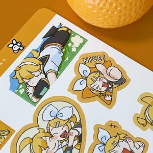 May include: Sheet of stickers featuring anime-style illustrations of a character with yellow hair and a blue bow. The stickers have a gold border and include the word "NICE!". An orange fruit sits in the background.