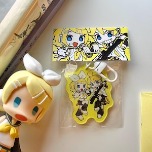 May include: A yellow acrylic keychain featuring a cartoon illustration of two anime-style characters, one holding a microphone and the other a keytar. The keychain is attached to a white plastic clip and includes a matching rectangular display card.