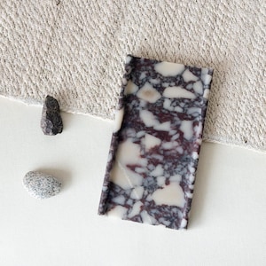 Puede incluir: Una bandeja rectangular con un diseño de terrazo en tonos burdeos, blanco y gris. Dos pequeñas piedras están al lado de la bandeja. La bandeja está sobre una superficie blanca con una tela texturizada en el fondo.