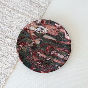 May include: A circular stone coaster featuring a polished surface with a marbled pattern of deep forest green, dark crimson red, and creamy white veins. The natural stone material displays a complex, swirling texture across the entire round face.