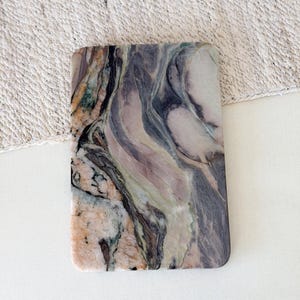 Puede incluir: Un posavasos rectangular de piedra con esquinas redondeadas. El posavasos presenta un patrón veteado natural en tonos grises, morados, verdes y beige. La piedra tiene un acabado pulido, y la estética general es orgánica y elegante.