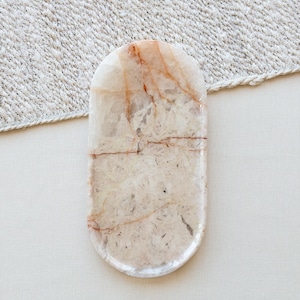 Puede incluir: Una bandeja de forma ovalada hecha de piedra pulida, con una base de color blanco roto translúcido y vetas irregulares de color marrón rojizo que recorren toda la superficie. La bandeja presenta un acabado liso y brillante con bordes redondeados.