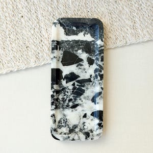 Puede incluir: Una bandeja rectangular con un patrón de mármol en blanco y negro de alto contraste con grandes fragmentos de piedra angulares. La superficie tiene un acabado pulido con vetas distintivas y formas variadas de piedra oscura y clara incrustadas.