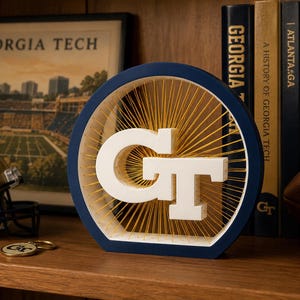 May include: A circular navy blue wooden frame containing a white Georgia Tech GT logo suspended by thin gold strings radiating from the center. The piece features a sunburst pattern created by the tension of the strings against the white letters.