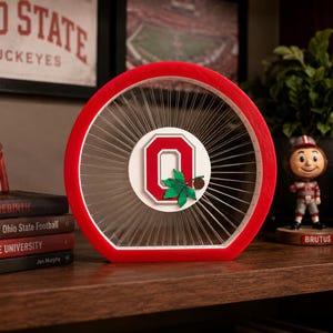 May include: A circular red wooden frame featuring a string art design with a white center disc displaying a red block O logo. A green buckeye leaf and a small brown buckeye nut are attached to the center of the logo, surrounded by thin white strings.