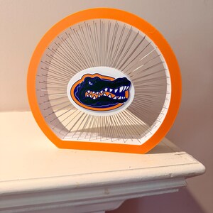 May include: A circular orange frame containing a dense web of thin silver metal pins radiating from the center. An oval sticker featuring a dark green alligator head with orange and blue accents is positioned in the middle of the pin array.