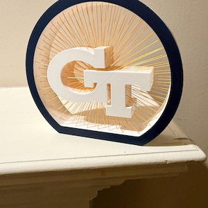 May include: A circular wall art piece featuring a dark blue outer frame and a white GT logo at the center. The logo is suspended within the frame by numerous thin, gold-colored radial strings that create a sunburst effect against the white interior.