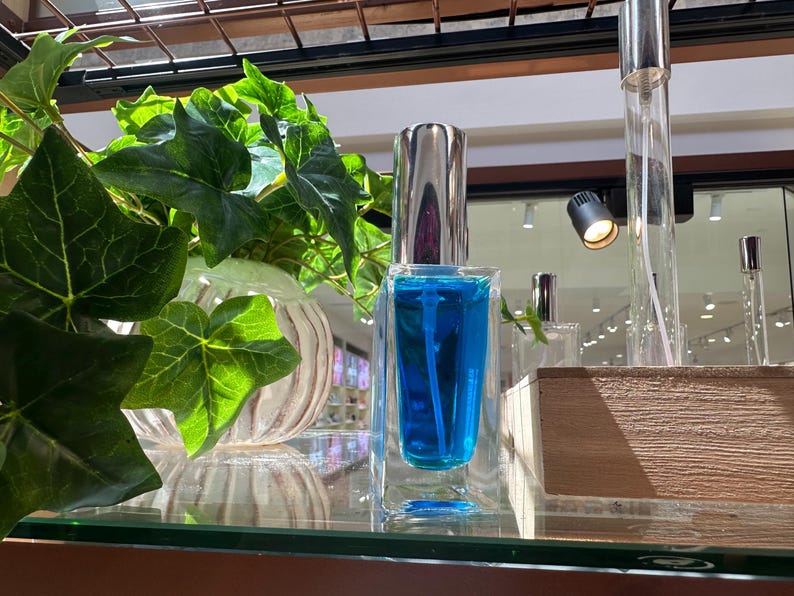May include: A clear glass perfume bottle with a silver cap, filled with bright blue liquid. The bottle is displayed on a glass shelf, with green foliage in the background. Other perfume bottles are also visible.