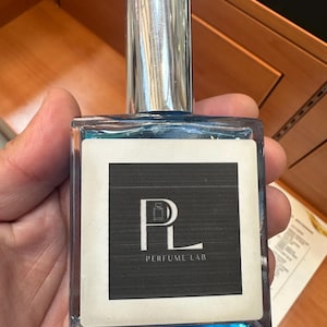May include: A rectangular perfume bottle with a silver cap and blue liquid. The label features the text "PL PERFUME LAB" in white on a black background. The bottle is held in a hand.