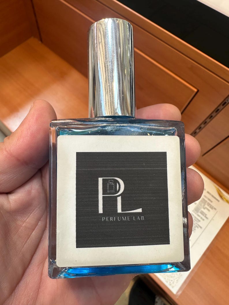 May include: A rectangular perfume bottle with a silver cap and blue liquid. The label features the text "PL PERFUME LAB" in white on a black background. The bottle is held in a hand.