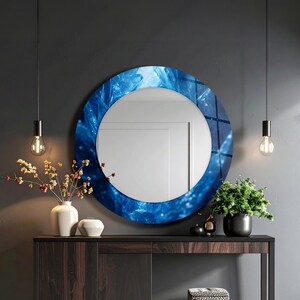 May include: A round wall mirror featuring a decorative frame with a vibrant blue abstract pattern resembling crystalline structures. The mirror has a smooth, reflective circular center and is mounted on a dark gray wall above a wooden console table.