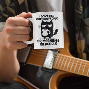 Puede incluir: Taza de cerámica blanca con el texto "I don't like morning people or mornings or people" y un gato de dibujos animados sosteniendo una taza de café. La taza tiene una ilustración de gato negro y texto negro.