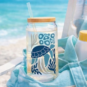May include: A clear glass tumbler with a bamboo lid and a straw. The tumbler features a blue sea turtle design with seaweed and bubbles. The tumbler is filled with water and sits in a blue beach bag, with the ocean in the background.