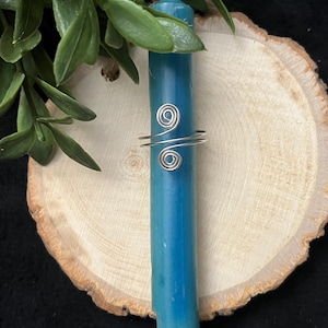 May include: A silver-colored ring with a spiral design is wrapped around a blue candle. The candle is resting on a wooden slice, with green leaves in the background. The candle is approximately 6 inches tall.