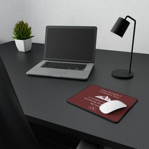 May include: A computer desk setup with a laptop, a burgundy mousepad with white text and a mountain graphic, a white mouse, a small potted plant, and a black desk lamp. The mousepad text reads: "they thought I climbed a hill..."