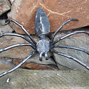 May include: A handcrafted metal spider sculpture with eight legs and a textured body. The spider is dark gray and appears to be made of forged metal. The sculpture is displayed against a backdrop of natural stone.